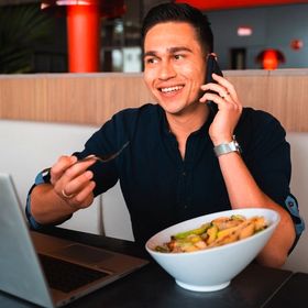 Young Asian male talks on the phone while eating a salad and looking at his laptop.