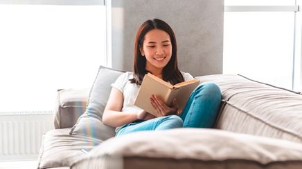 Woman in white T-shirt and denim pants reading a book while lying on a sofa.
