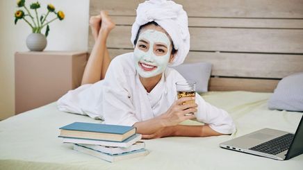 Asian woman in a robe wearing a mud mask while relaxing on a bed with books and a drink.
