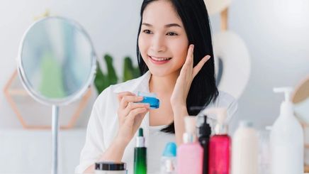 Asian woman applying skincare products in front of mirror.