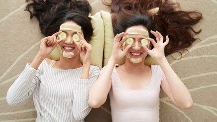 Two women wearing cucumber face masks lying on a carpet.