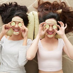 Two women wearing cucumber face masks lying on a carpet.
