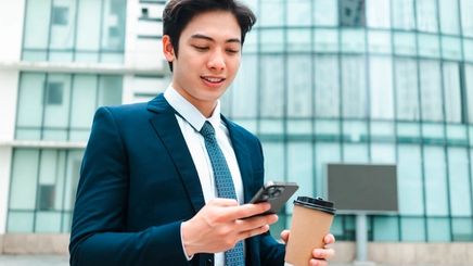 Young Asian man in a blue suit holding a phone and a cup of coffee.
