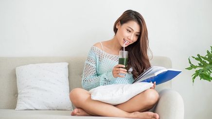 Asian woman sitting on sofa, reading a book while sipping green drink