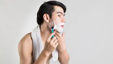 A man applies shaving cream on his face while staring into a bathroom mirror.