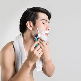 A man applies shaving cream on his face while staring into a bathroom mirror.