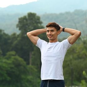 Young Asian male in white shirt stretches his arms behind his head at a park. 