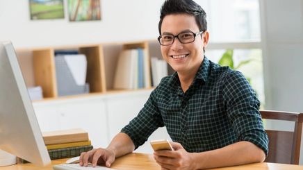 A smiling man holds a phone while using a computer.