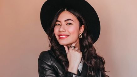 Woman wearing black leather jacket and wide-brimmed hat smiling to the camera.