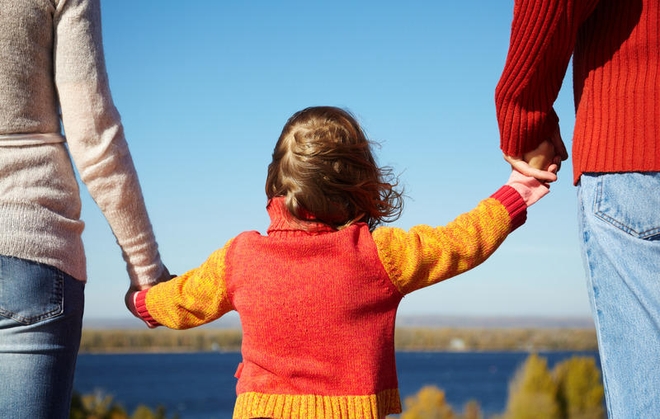 Liten jente blir leid i hver sin hånd av mamma og pappa. Fotografert utendørs, bakfra.