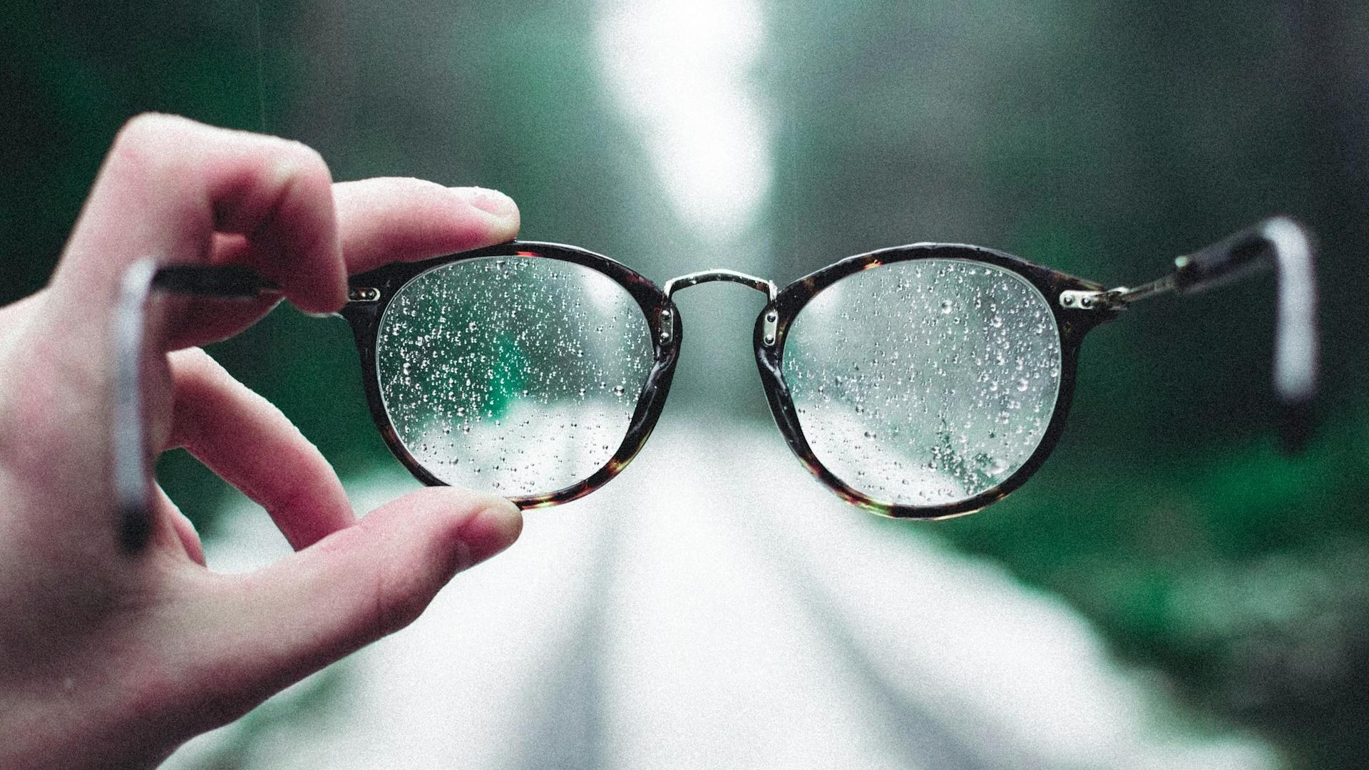 Mains tenant une paire de lunettes avec des gouttelettes d'eau, illustrant la manipulation et le contrôle des verres dans l'atelier de l'opticien.