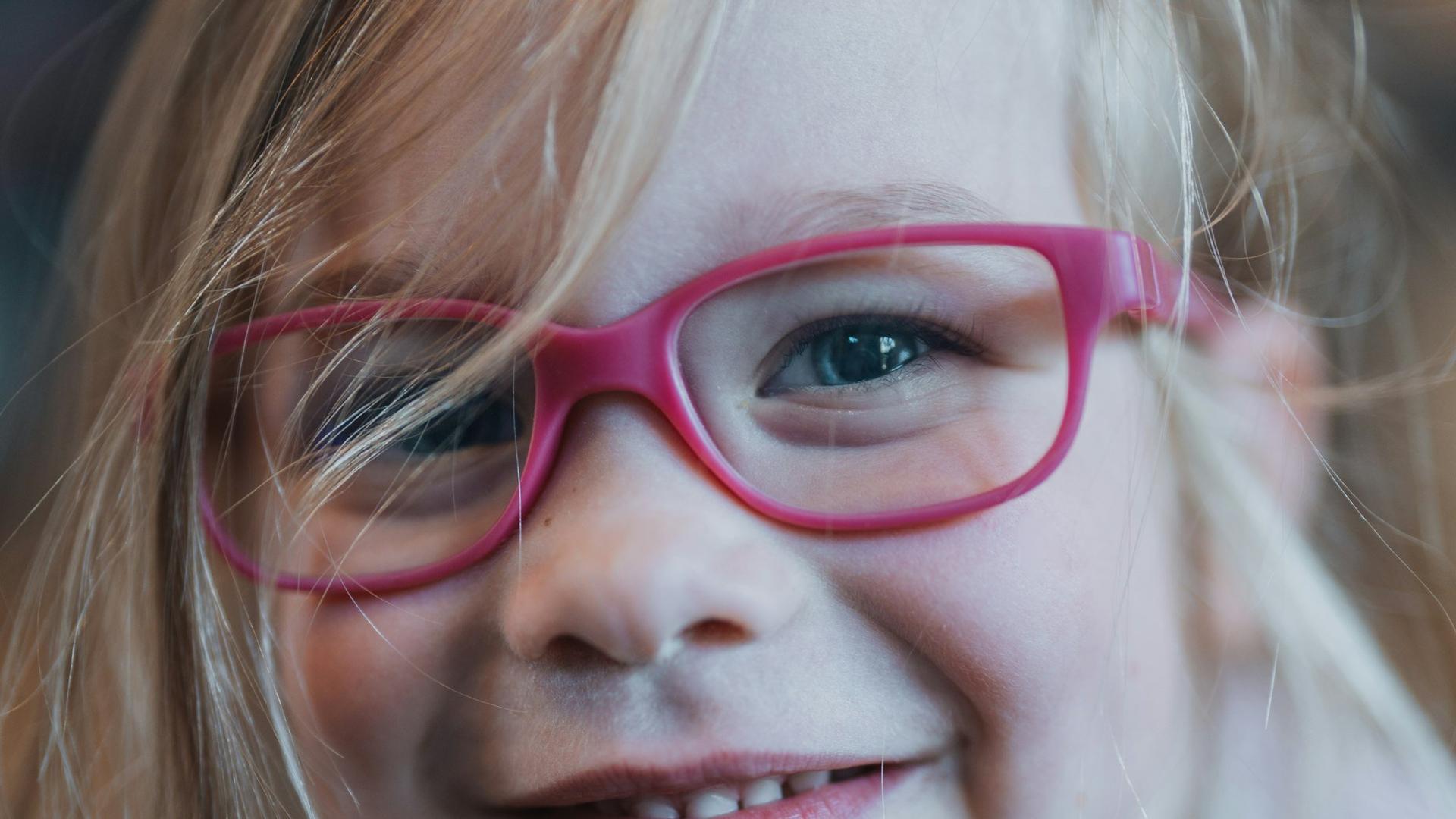 Petite fille souriante portant des lunettes roses, le port de lunettes étant le premier traitement de l'amblyopie chez l'enfant