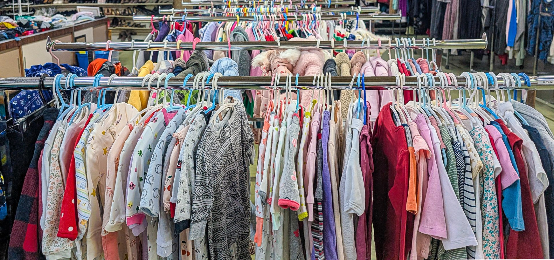 Cette photographie montre une rangée de vêtements colorés suspendus sur des cintres métalliques dans une friperie. Les articles, allant des t-shirts aux manteaux, sont enlignés.