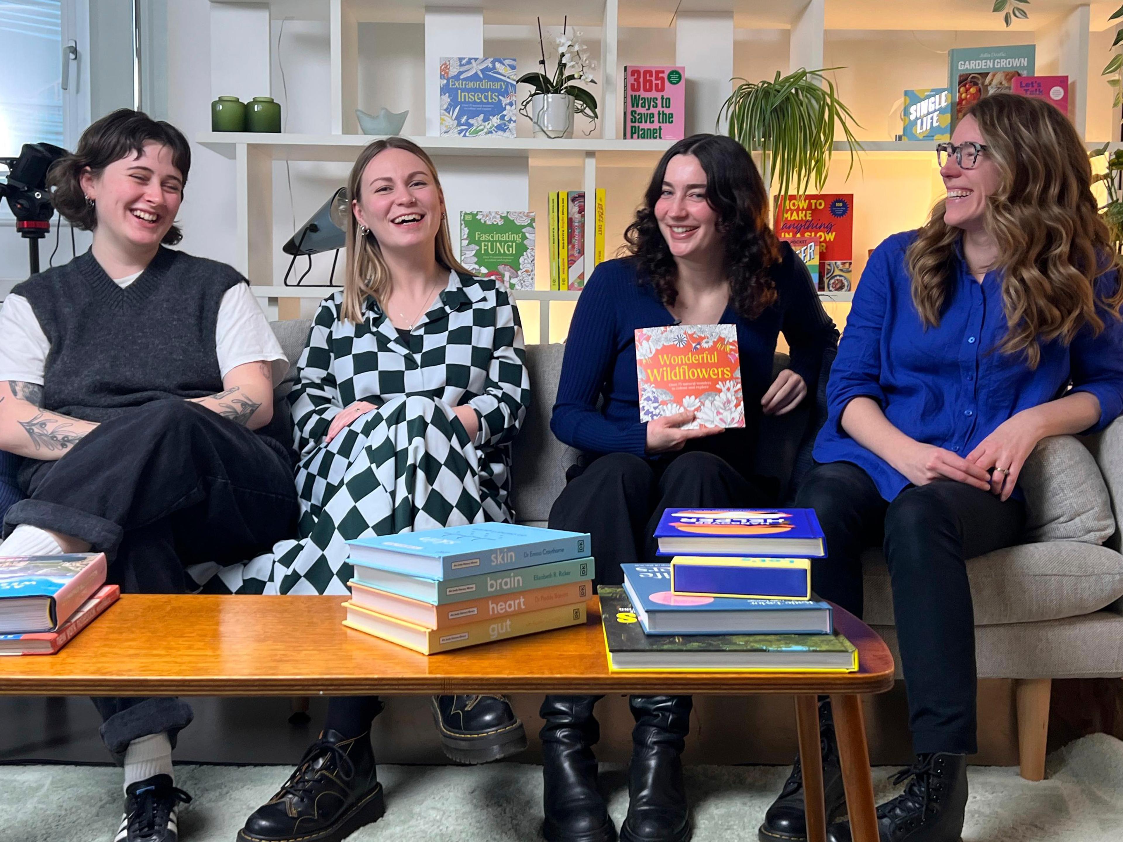Four women seated on a sofa in a studio setting, smiling and chatting, with stacks of books on a coffee table and bookshelves behind them.