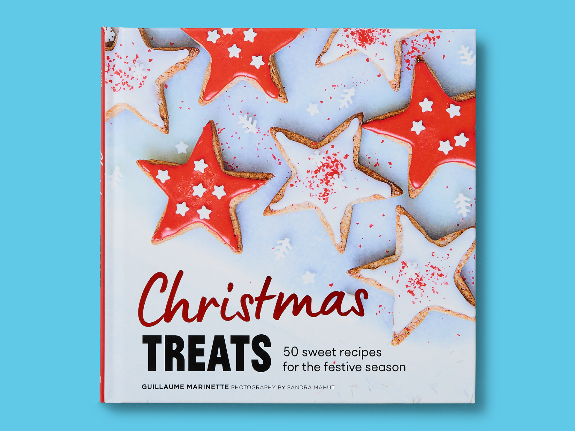 Book titled "Christmas Treats" on a bright blue background, featuring iced star-shaped biscuits in red and white scattered across the cover with festive sprinkles. Handwritten-style title appears above bold subtitle reading "50 sweet recipes for the festive season," with author Guillaume Marinette and photography credit to Sandra Mahut at the bottom.