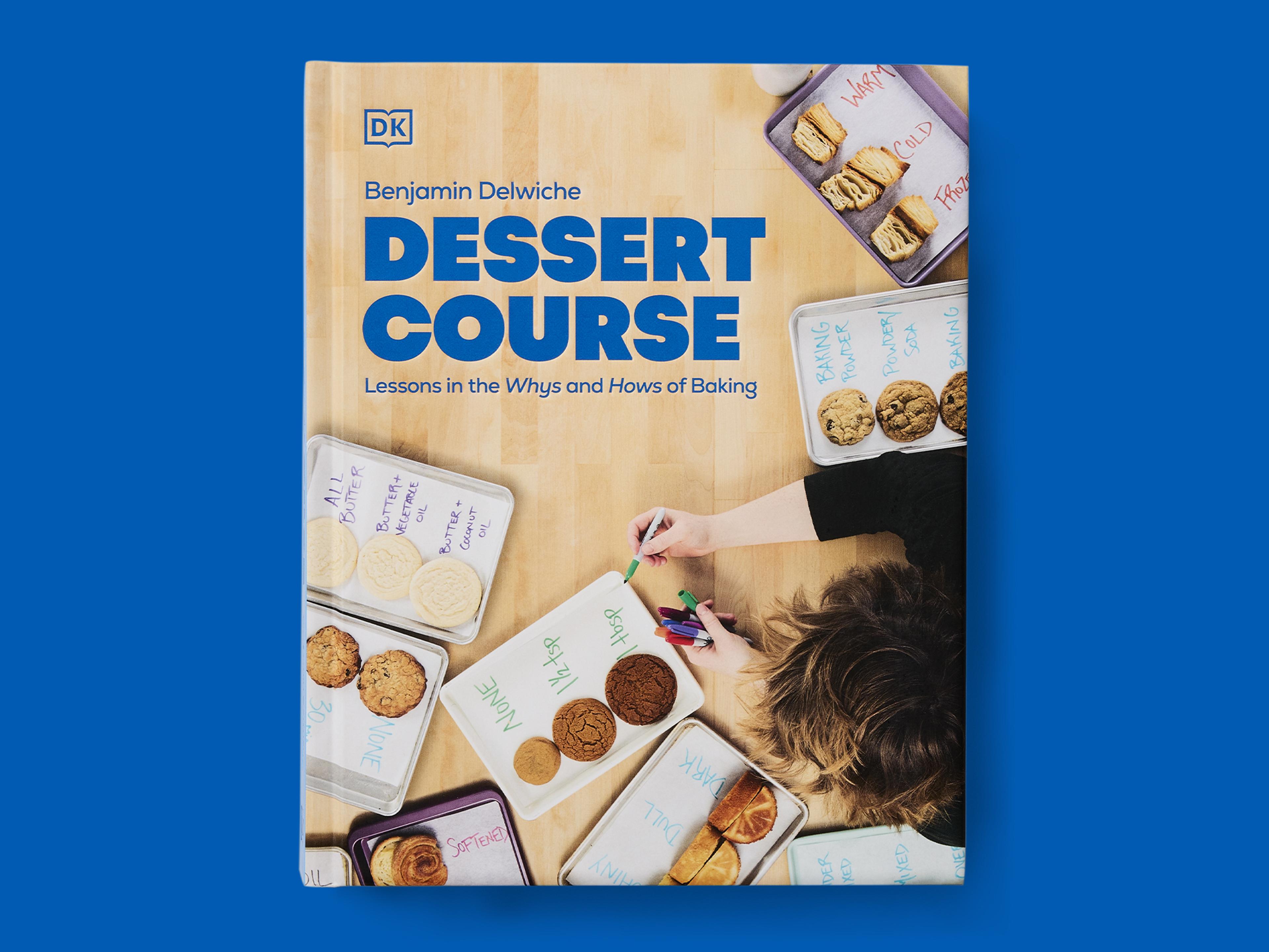 Book cover titled "Dessert Course: Lessons in the Whys and Hows of Baking" by Benjamin Delwiche, featuring an overhead photograph of baking trays with labelled cookies and pastries, hands holding coloured pens, and the DK logo, set against a blue background.