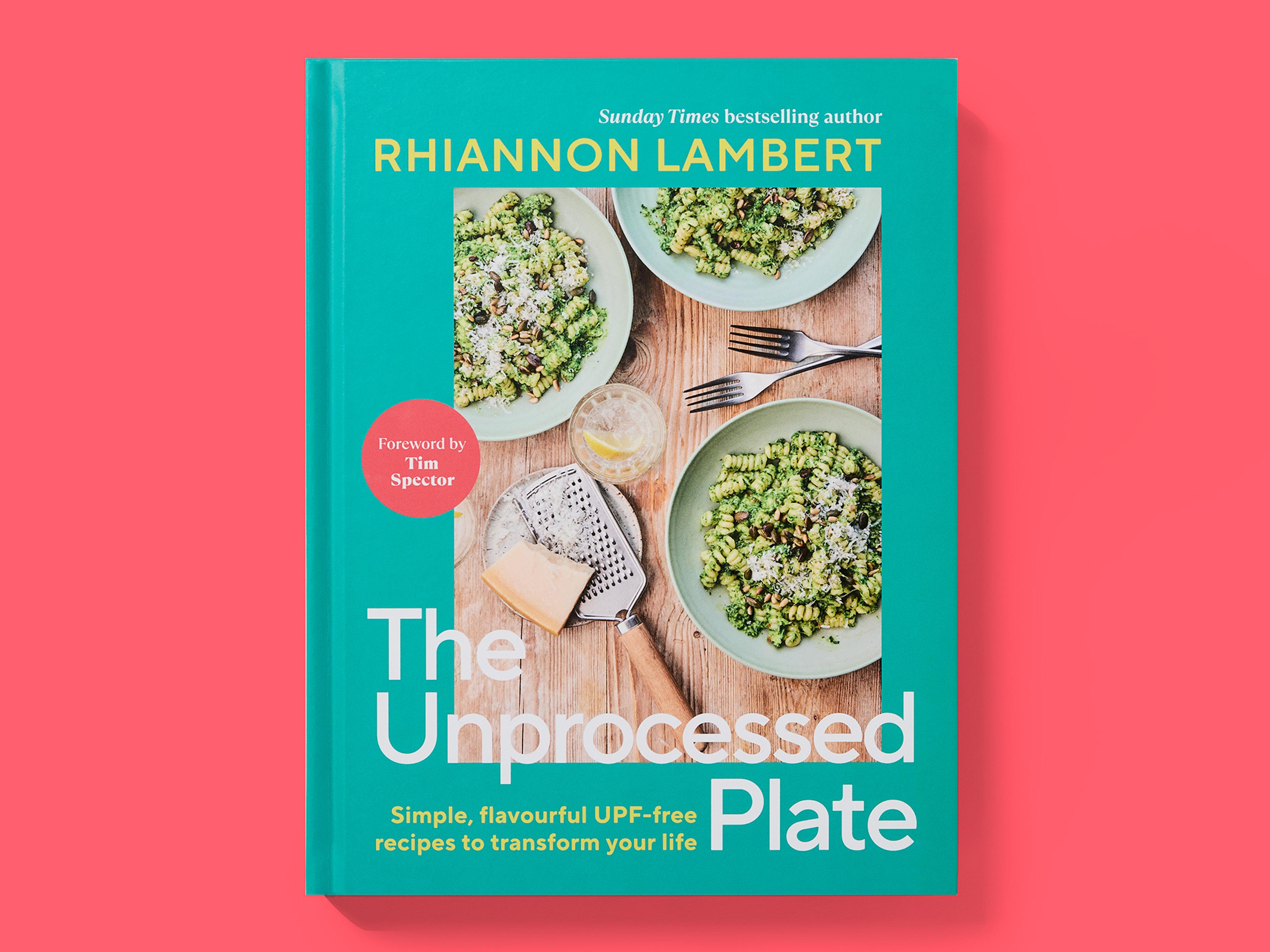 Teal book cover titled "The Unprocessed Plate" by Rhiannon Lambert, featuring a food photograph of pasta dishes, forks, cheese, and a grater on a wooden table, with the subtitle "Simple, flavourful UPF-free recipes to transform your life" and a red badge noting a foreword by Tim Spector, set against a pink background.