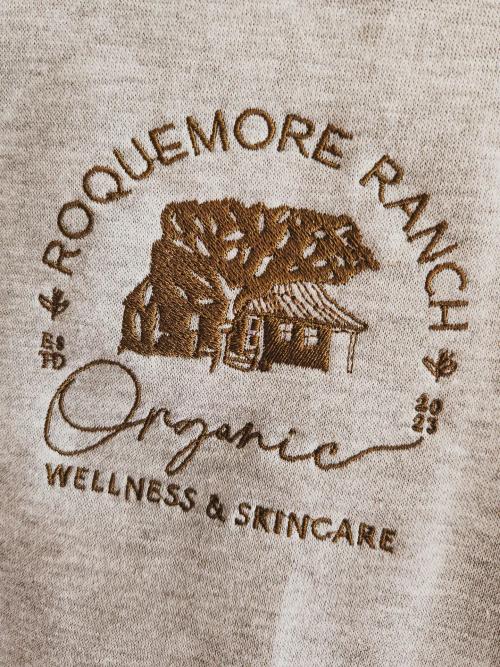 "Roquemore Ranch Organic Wellness & Skincare" circular farmhouse logo embroidered on a heather oatmeal sweatshirt