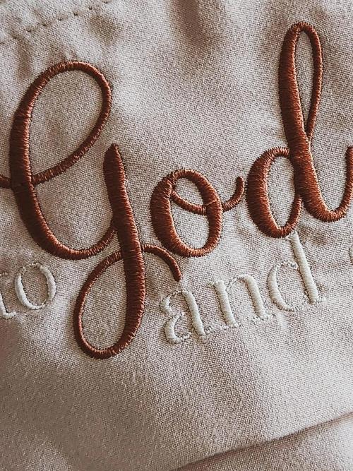 Close-up of natural linen fabric embroidered with cursive script reading "Grand..." in rust-brown thread, part of a larger phrase