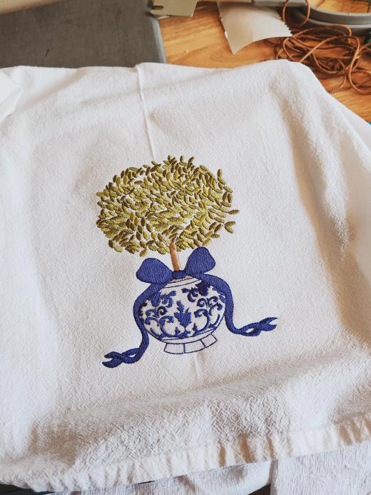 White flour sack kitchen towel embroidered with a chinoiserie-style topiary tree in a blue-and-white ginger jar tied with a navy bow
