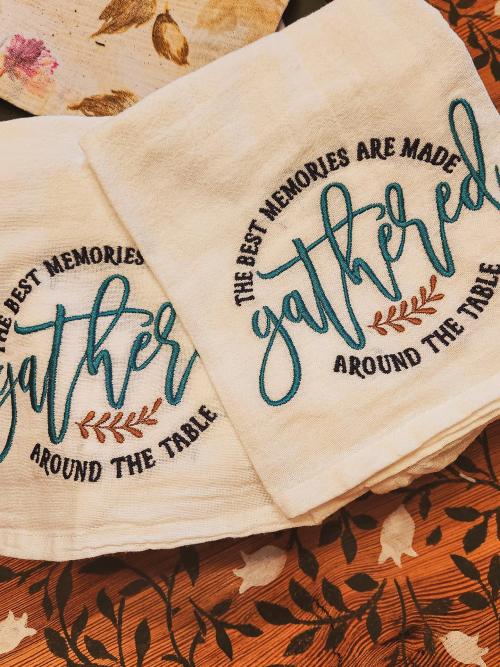 Matching flour sack kitchen towels embroidered with "The best memories are made gathered around the table" in teal script with rust leaf accents