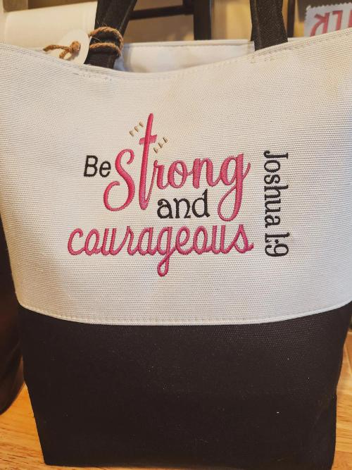 Black and white canvas tote bag embroidered with "Be Strong and Courageous, Joshua 1:9" in pink and black lettering with a small gold cross