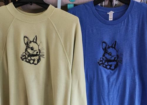 Tan sweatshirt and heather blue adult t-shirt hanging side by side, each embroidered with the same bunny-head-and-flowers design in a single thread color.