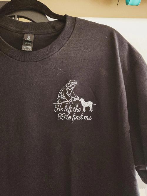 Black t-shirt with a white line-art embroidered shepherd kneeling beside a lamb and the text "He left the 99 to find me"