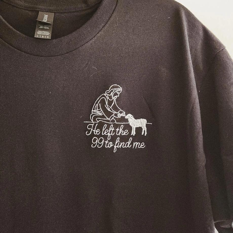 Black t-shirt with a white line-art embroidered shepherd kneeling beside a lamb and the text "He left the 99 to find me"