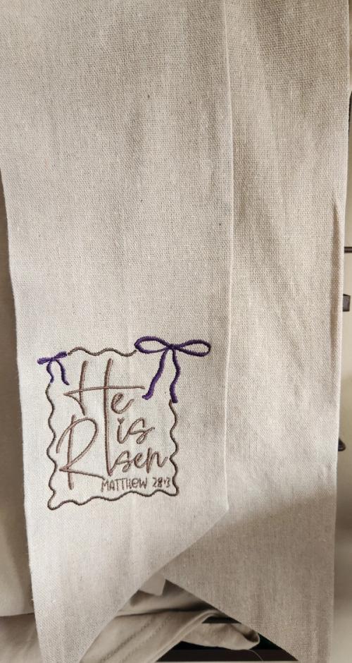 Cream linen ribbon sash embroidered with "He is Risen, Matthew 28:6" inside a scalloped frame topped with a purple bow.