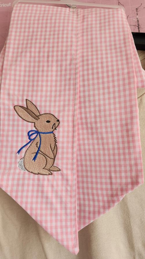 Pink and white gingham wreath sash embroidered with a sitting rabbit wearing a blue ribbon bow around its neck.