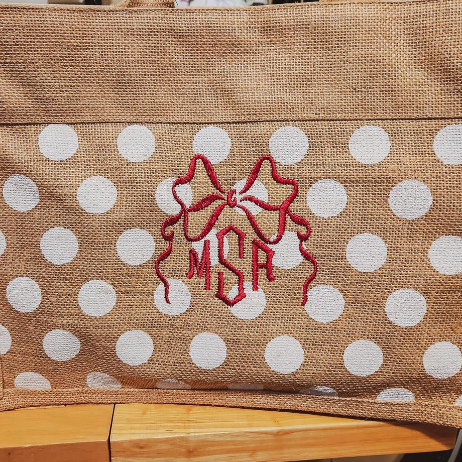 Three-letter red script monogram with bow and star ribbon design on a burlap tote bag with white polka dots