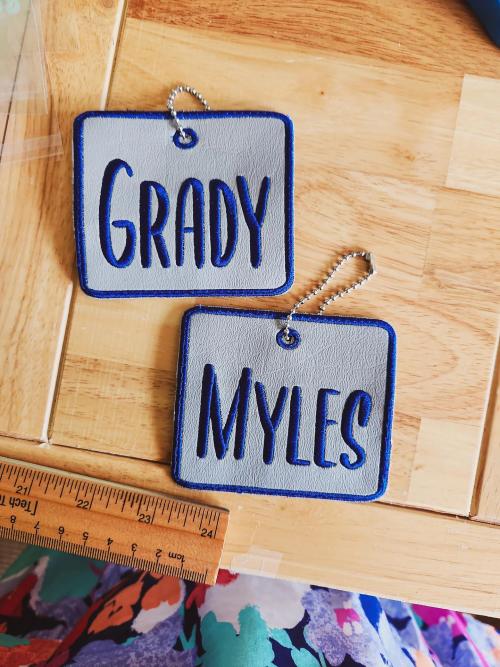 Two gray vinyl bag tags on ball-chain key rings embroidered with the names "Grady" and "Myles" in navy block letters