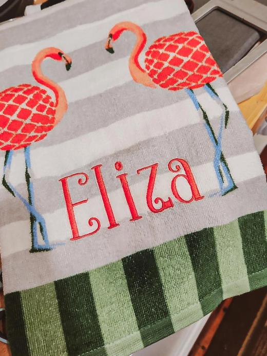 Red script name "Eliza" embroidered below two flamingo appliques on a gray-striped hand towel with a green buffalo check border