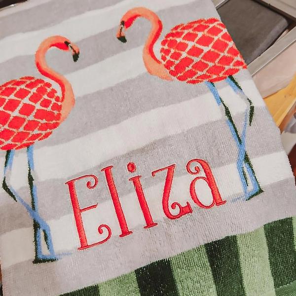 Red script name "Eliza" embroidered below two flamingo appliques on a gray-striped hand towel with a green buffalo check border