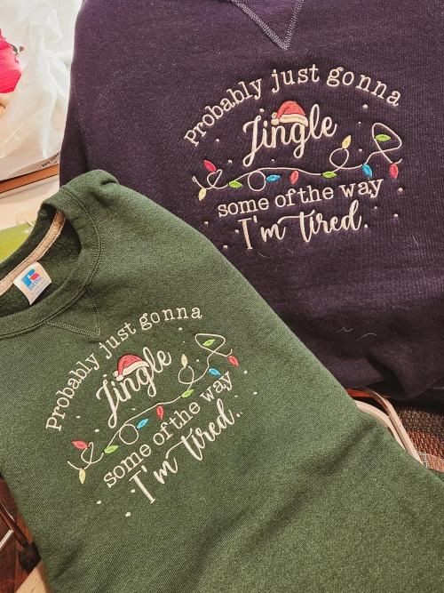 Purple and green pullover sweatshirts embroidered with "Probably just gonna Jingle some of the way I'm tired" and a Santa hat with a string of colorful Christmas lights