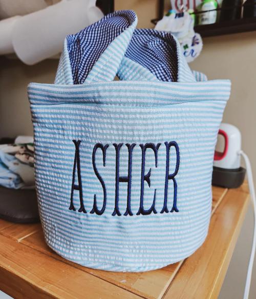 Navy block name "Asher" embroidered on a light blue seersucker fabric storage bucket with navy gingham lining