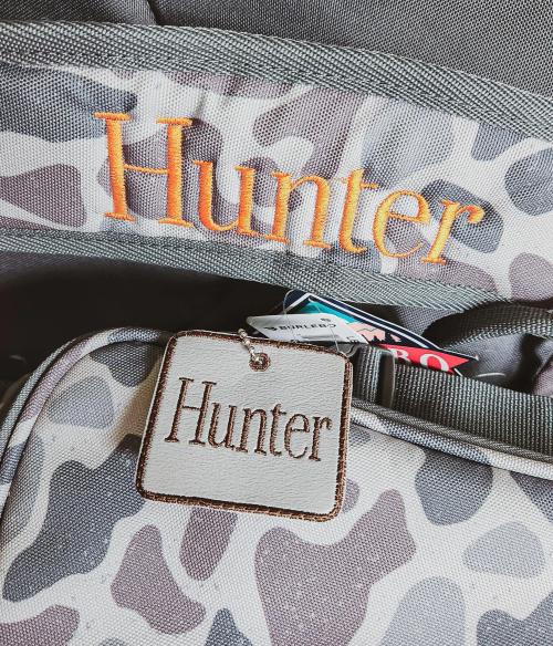Orange script name "Hunter" embroidered on a gray camo duffel bag strap, with a matching leather-look "Hunter" luggage tag attached