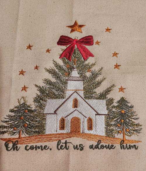 Close-up of a canvas wall hanging embroidered with a white church, Christmas trees, gold stars, and a red bow above the phrase "Oh come, let us adore Him"