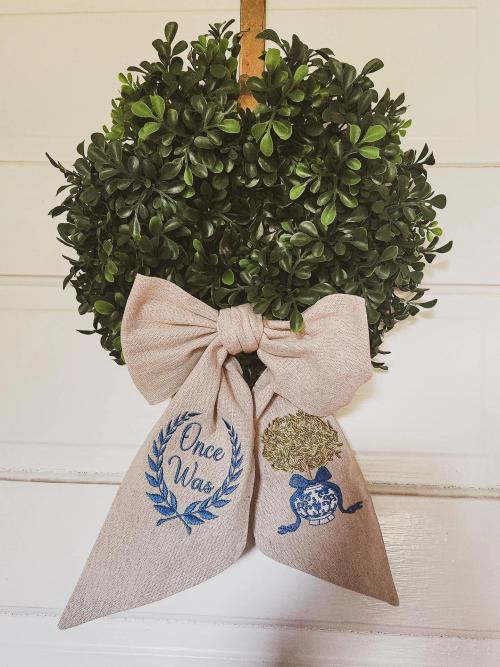 Boxwood wreath with a linen bow embroidered with the words "Once Was" inside a leaf wreath and a blue-and-white ginger jar topiary motif on the ribbon tails.