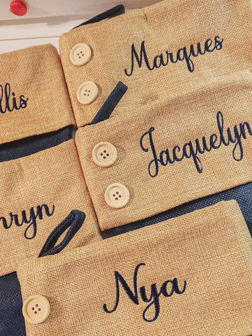 Stack of burlap drawstring gift bags each embroidered in navy script with a different first name, including Marques, Jacquelyn, and Nya