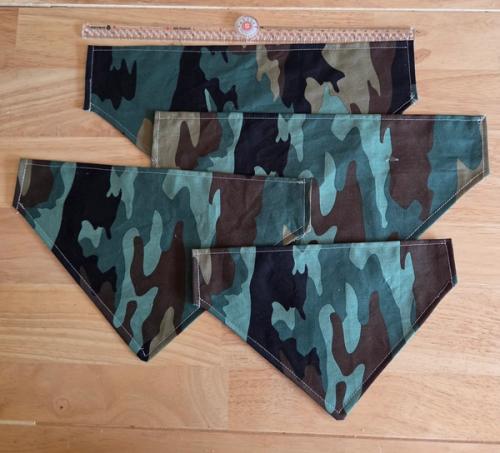 Four camouflage-print, slide-on pet bandanas laid out on a wood floor next to a ruler for size reference.
