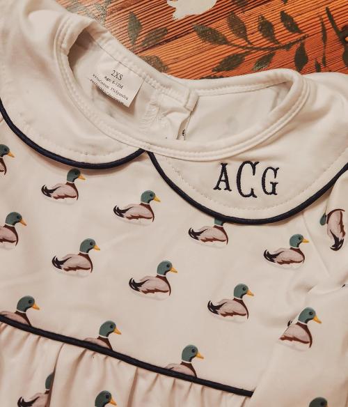 Navy block three-letter monogram "ACG" on the collar of a cream infant dress with a mallard duck print and navy piping