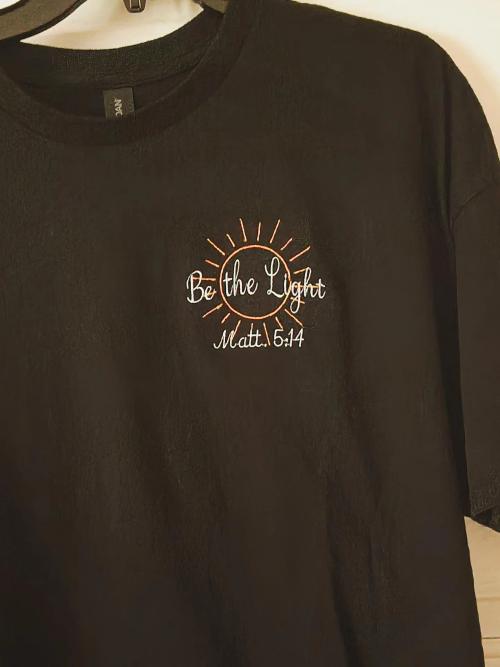 Black t-shirt embroidered with "Be the Light, Matt. 5:14" inside an orange sunburst design