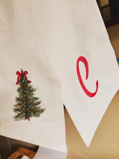Corner of a cream linen towel embroidered with a decorated Christmas tree and a separate script monogram letter "C" in red thread.