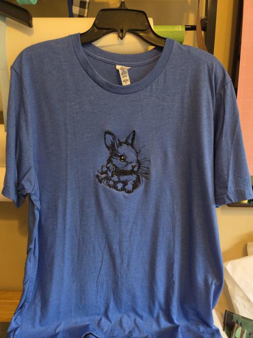 Heather blue adult t-shirt embroidered with a detailed line-art bunny head holding a small flower bouquet, centered on the chest.