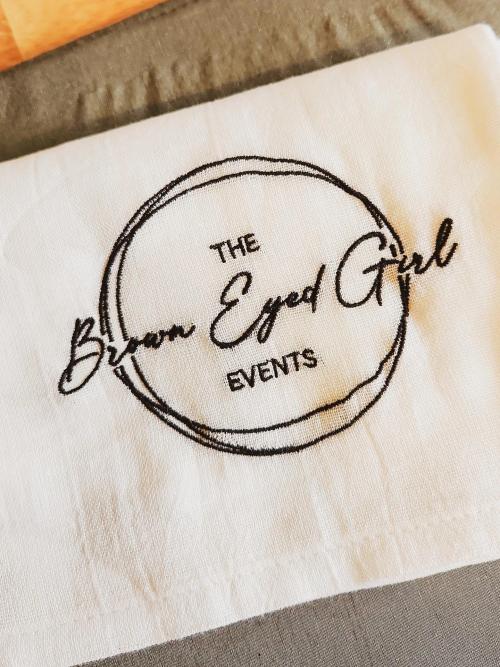 "The Brown Eyed Girl Events" circular business logo embroidered on a folded white cloth napkin