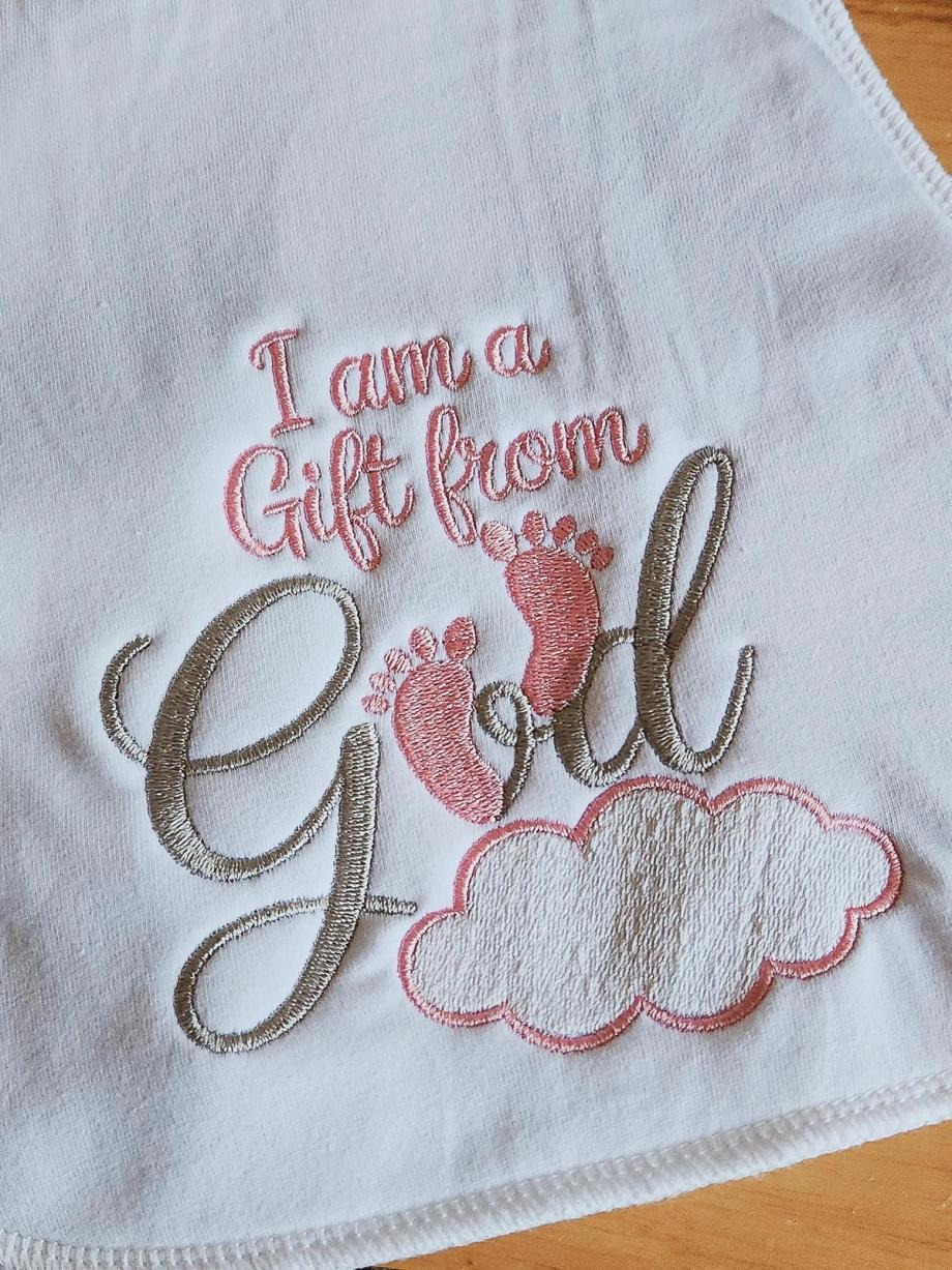 White baby burp cloth embroidered with "I am a Gift from God" in script and pink appliqué baby footprints resting on an embroidered cloud