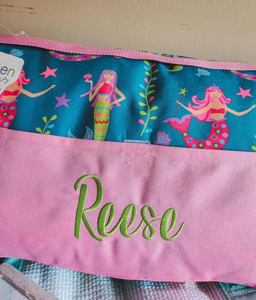 Green script name "Reese" embroidered on the pink panel of a kids' bag with a mermaid-print lining
