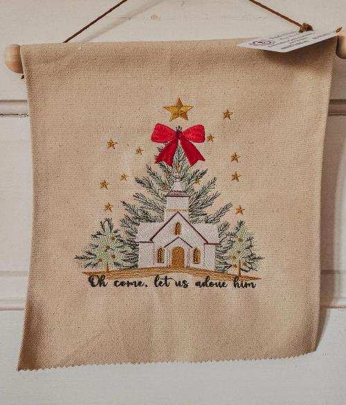 Canvas wall hanging embroidered with a white church, Christmas trees, gold stars, and a red bow above the phrase "Oh come, let us adore Him"